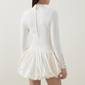 Model wearing Aje cream dress with ribbed long-sleeve top, gold button details on cuffs, and voluminous bubble-hem skirt, shown from back