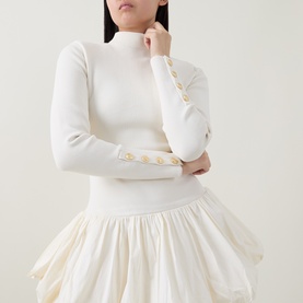 Model wearing Aje cream dress with ribbed mock-neck top, gold button cuff detail, and voluminous skirt hem.