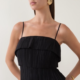 Model wearing Aje black pleated dress with tiered ruffle bodice and thin shoulder straps, styled with gold hoop earrings and bracelet