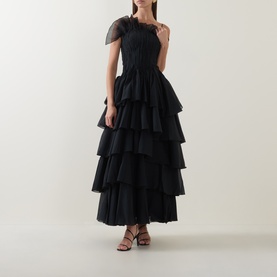 Model wearing Aje black tiered tulle maxi dress with ruched one-shoulder bodice and sheer cap sleeve, paired with red heeled sandals