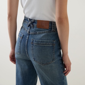 Model wearing Aje jeans in Stormy Wash, close-up of back view showing leather logo patch and classic five-pocket design, styled with white tee.