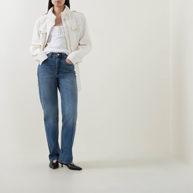 Model wearing Aje jeans in Stormy Wash, a mid-blue straight-leg denim style, paired with a cream jacket and white top.
