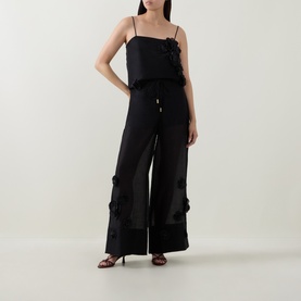 Model wearing Aje black sleeveless camisole top with floral appliqu&eacute; detail, styled with matching wide-leg black trousers and heeled sandals.