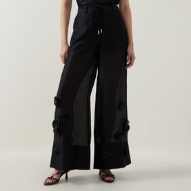 Model wearing Aje black wide-leg trousers with floral appliqu&eacute; detail, styled with brown strappy heeled sandals.