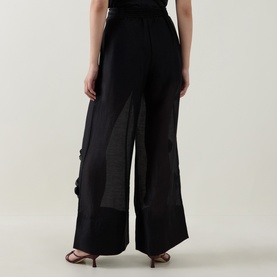 Model wearing Aje black wide-leg trousers with sheer panel detail and tie waist, styled with strappy heeled sandals, rear view.