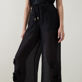 Model wearing Aje black wide-leg trousers with floral appliqu&eacute; detail and drawstring waist in sheer lyocell-linen blend.