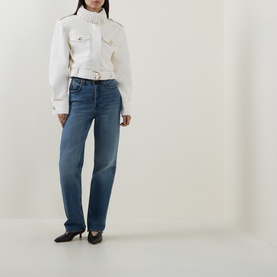 Model wearing Aje Mossan Utility Cropped Jacket in ivory with gold hardware, styled with blue straight-leg jeans and black kitten heels.