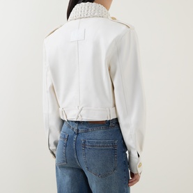 Model wearing Aje Mossan Utility Cropped Jacket in ivory, back view showing braid collar, gold-toned hardware and epaulettes, styled with blue jeans