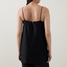 Model wearing Aje black cami top, back view, thin straps with gold clip detail, relaxed longline silhouette in linen-lyocell blend.