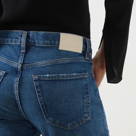 Model wearing Citizens of Humanity Slim Jeans in cruise mid-blue denim, close-up of back pockets with gold-tone logo plaque detail.