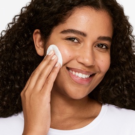 Model with Clinique exfoliator, shade 3 Combination/Oily, applying cotton pad to cheek, wearing white top