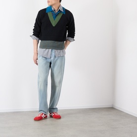 Model wearing Dries Van Noten black wool V-neck knit with green chevron detail, styled with check shirt, light wash jeans and red trainers.