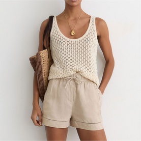 Model wearing Reiss beige linen drawstring shorts, styled with cream crochet knit vest top and gold pendant necklace, carrying woven bag.