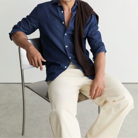 Model wearing Reiss dark blue wash shirt, unbuttoned at collar, styled with cream trousers and brown jumper over shoulders