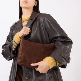 Model carrying Miu Miu Camoscio Small Shoulder Bag in Moro brown suede with embossed logo, styled with dark leather coat and yellow knit sweater.