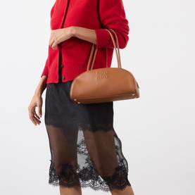 Model carrying Miu Miu Beau Softy Logo Bag in cognac leather with gold-toned hardware and top handles. Styled with red knitwear and black lace skirt.