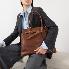 Model carrying Miu Miu Aventure Medium Leather Tote Bag in Rovere with gold-tone hardware and top handles, styled with grey blazer and blue shirt.