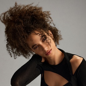 Model with voluminous, styled curly hair, shot to showcase Oribe Gold Lust Dry Heat Protection Spray results, wearing a black top.