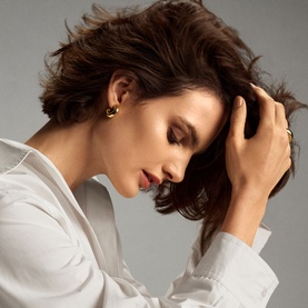 Model with hands through tousled dark hair, styled using Oribe Gold Lust Dry Heat Protection Spray, wearing white shirt