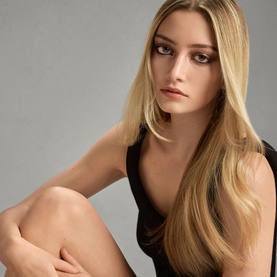 Model with Oribe Gold Lust Dry Heat Protection Spray, showcasing sleek, glossy long blonde hair styled straight against a grey background.