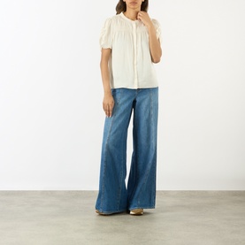 Model wearing Paige Antique White short-sleeve top with pleated detail, styled with wide-leg blue denim jeans and gold flat shoes.