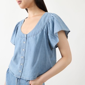 Model wearing Paige top in Palos Verdes blue denim with flutter sleeves, scoop neck, and button-front detail, styled with matching wide-leg trousers.