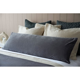 Belledorm Velvet Bolster Cushion in charcoal, styled on a bed with cream and teal cushions against a panelled headboard.
