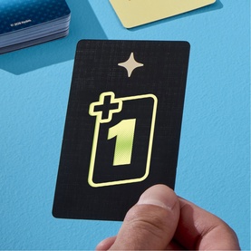 Black playing card with gold star and neon green +1 symbol, held over blue surface with additional cards visible nearby.