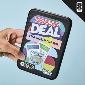 Monopoly Deal FIFA World Cup card game in black tin, held in hand, showing colourful game cards and multi-colour design