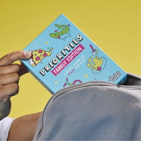 Priorities Family Edition card game in multi-coloured box, held above a grey backpack against a yellow background