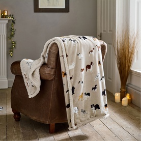 Deyongs oatmeal throw with multicoloured dog print, sherpa lining visible, draped over a brown leather armchair in a cosy living room setting.