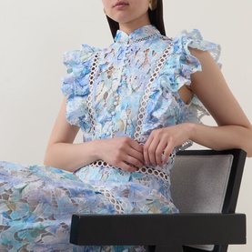 Model wearing Elliatt dress in bluemulti, featuring floral lace panels, ruffle sleeves, and high neck, seated on a dark chair.