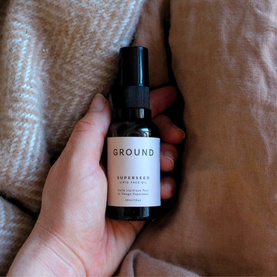 Ground Wellbeing Superseed Lipid Face Oil in a dark glass pump bottle, held in hand against a soft beige textured fabric background.