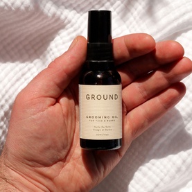 Ground Grooming Oil for Face & Beard in dark glass pump bottle with cream label, held in hand against white fabric background