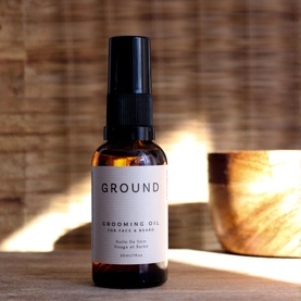 Ground Grooming Oil for Face & Beard in amber glass spray bottle with black pump, styled on wooden surface with bowl in background.