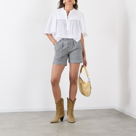 Model wearing Isabel Marant Etoile Douna Embroidered Blouse in white with grey shorts and tan cowboy boots, carrying a beige shoulder bag.