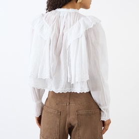 Model wearing Isabel Marant Etoile white cotton blouse with tiered ruffle detail, lace trim hem, and long sleeves, styled with tan trousers