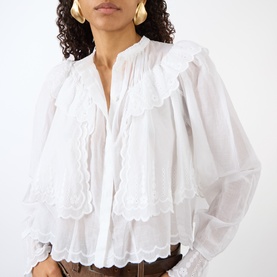 Model wearing Isabel Marant Etoile white cotton blouse with scalloped ruffle bib, bishop sleeves, and V-neckline, styled with tan trousers.