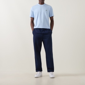 Model wearing Lacoste Croc Logo T-Shirt in Rill blue, crew neck, short sleeves, styled with navy trousers and white trainers.
