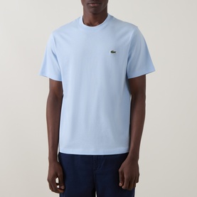 Model wearing Lacoste Croc Logo T-Shirt in Rill blue, crew neck, short sleeves, small croc logo on chest, styled with navy trousers.