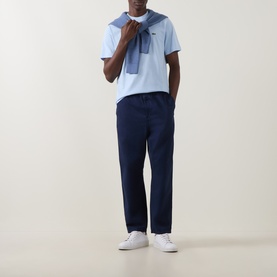 Model wearing Lacoste Croc Logo T-Shirt in Rill blue, styled with a blue sweater over shoulders, navy trousers and white trainers.
