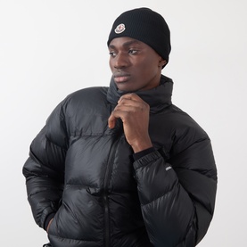 Model with Moncler Ribbed Logo Beanie Hat in black, ribbed knit with logo patch, styled with a black quilted puffer jacket.
