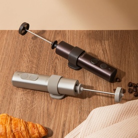 Two handheld electric milk frothers in dark brown and silver, shown on a wooden surface with croissant and coffee beans