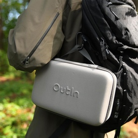 OutIn Nano Portable Espresso Machine in grey hard-shell carry case with embossed logo, clipped to black hiking backpack outdoors
