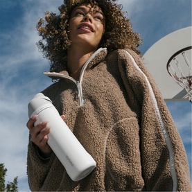 Model carrying a white insulated water bottle, styled with a tan sherpa fleece half-zip, outdoors near a basketball hoop