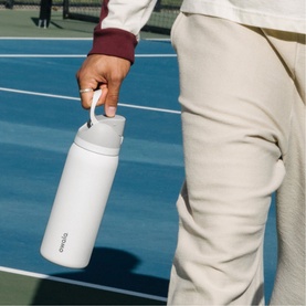 Model with white insulated water bottle with carry handle, held at a tennis court, styled with cream trousers and burgundy hoodie