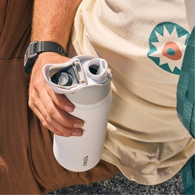 White insulated travel mug with open flip lid held in hand, interior compartment visible, casual outdoor setting