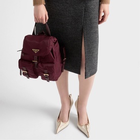 Model carrying small burgundy nylon backpack with gold hardware and buckle straps, styled with a grey midi skirt and gold pointed-toe heels.