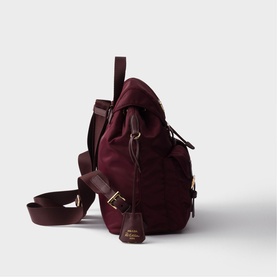 Burgundy canvas backpack with dark brown leather trim, gold-tone hardware, adjustable shoulder strap, and branded charm detail