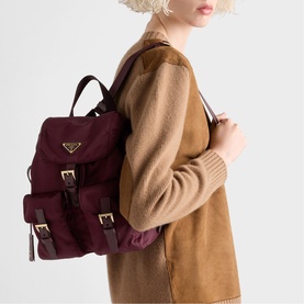 Model carrying burgundy Prada nylon backpack with gold-tone hardware and buckle straps, styled with a camel coat